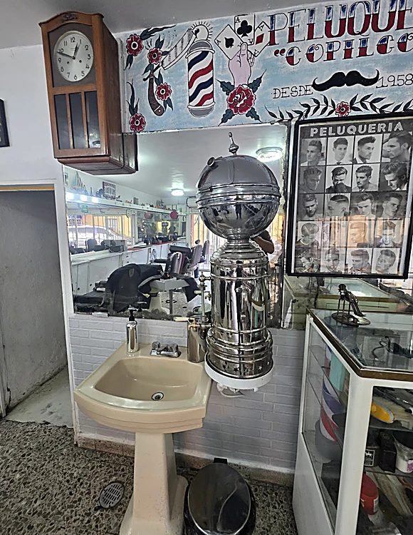 Rincón del fondo de la peluquería: reloj de péndulo en caja de madera, mural pintado con rosas, poste y las palabras Peluquería Copilco desde 1958, esterilizador cromado en forma de globo, lavabo color hueso y vitrina