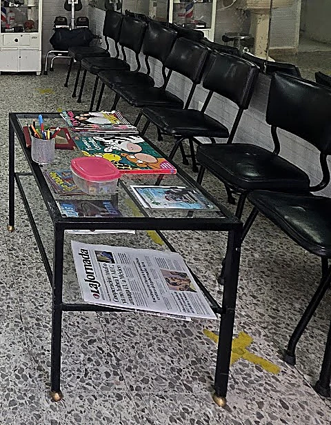 Mesa de vidrio con revistas, lápices y un periódico, junto a la fila de sillas negras de la espera