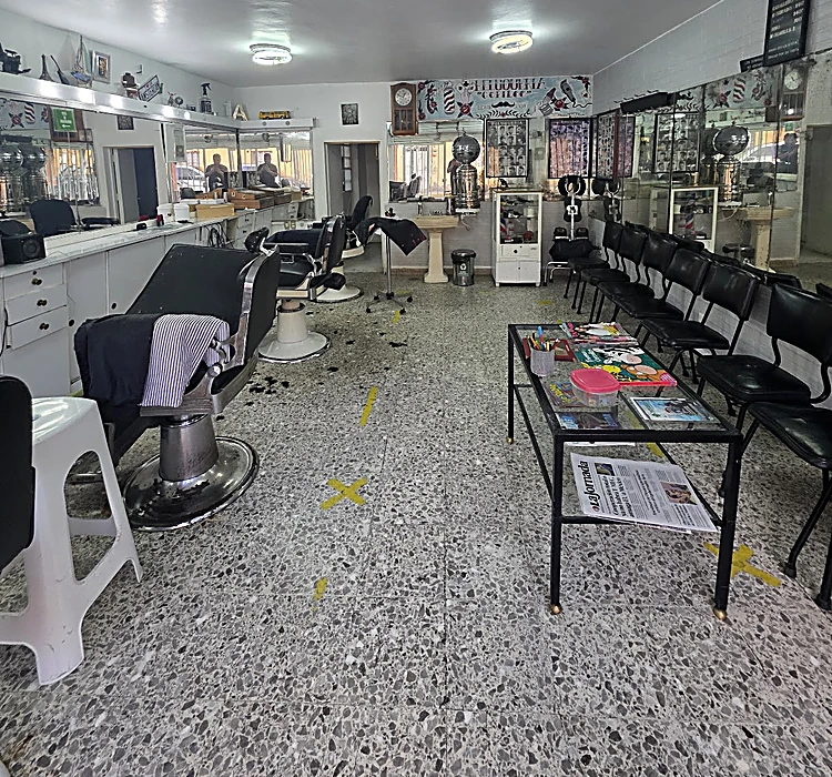 El salón completo: espejos corridos con gabinetes blancos a la izquierda, sillones de peluquería, una mesa de vidrio con revistas y periódico y una fila de sillas negras para esperar turno a la derecha